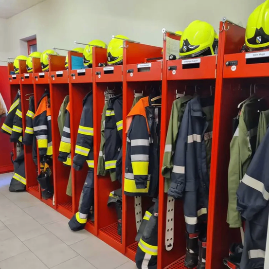 Eine Reihe roter Schließfächer mit Feuerwehrausrüstung, einschließlich Helmen, Jacken und Hosen. Die Schließfächer haben weiße Etiketten und sind ordentlich organisiert. Der Raum hat eine weiße Decke und einen gefliesten Boden.