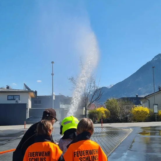Drei Menschen in Feuerwehruniformen stehen vor einem Springbrunnen mit aufspritzendem Wasser. Sie sind wahrscheinlich Feuerwehrleute. Dahinter befindet sich ein Berg.