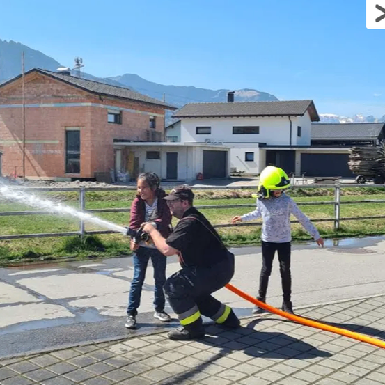 Ein Feuerwehrmann benutzt einen Wasserschlauch, während zwei Kinder an einem sonnigen Tag vor einem Haus zusehen.