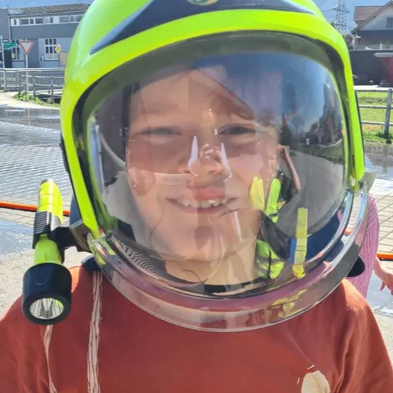Ein junger Junge in einem roten T-Shirt trägt einen gelben Helm mit reflektierenden Streifen, einem Logo und einer Taschenlampe. Er steht an einem Außenbereich mit Gebäuden und Bergen im Hintergrund.