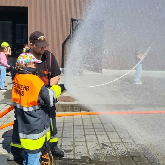 Ein Feuerwehrmann und ein Kind spielen mit einem Wasserschlauch vor einem Gebäude mit Bergen im Hintergrund.