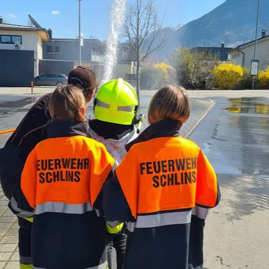 Eine Gruppe von Personen in Sicherheitsjacken und Helmen steht vor einem Hydranten. Einer trägt einen gelben Helm. Hinter ihnen spritzt ein Wasserbrunnen Wasser. Im Hintergrund befinden sich ein Gebäude, ein Berg und ein Auto.