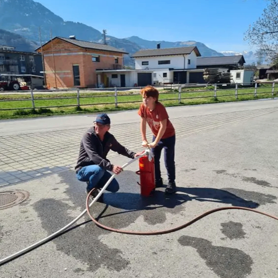 Zwei Personen sind auf einer Straße, eine hält einen Schlauch, der an einem Feuerlöscher angeschlossen ist. Die Umgebung hat Häuser und Berge.