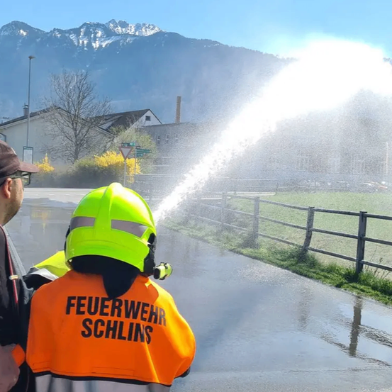Ein Feuerwehrmann in einem neongrünen Helm sprüht Wasser auf eine nasse Straße, während ein Mann in der Nähe steht. Dahinter befindet sich ein Zaun, Gebäude und Berge.