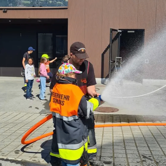 Ein Feuerwehrmann und ein Kind benutzen einen Wasserschlauch auf einem Bürgersteig. Hinter ihnen beobachten eine Gruppe von Kindern und ein Mann.