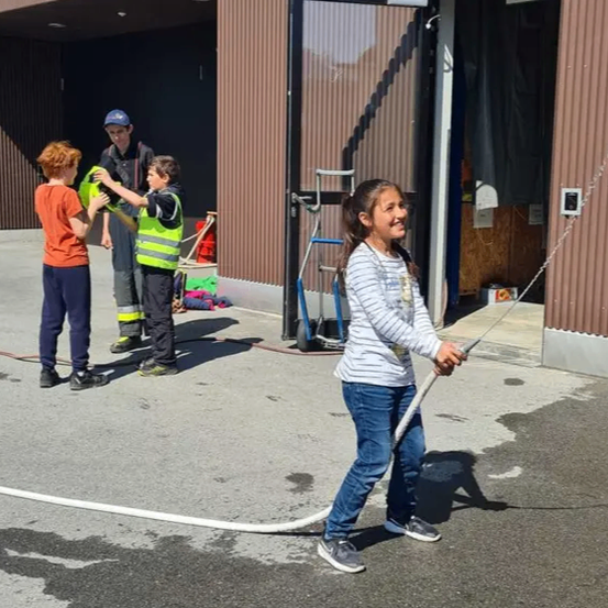 Ein junges Mädchen hält einen Wasserschlauch, während ein Junge und ein Mann eine gelbe Weste halten. Sie stehen vor einem Gebäude mit offener Tür.