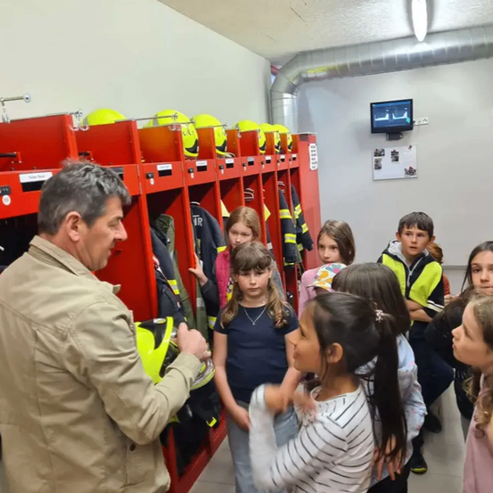 Ein Mann spricht mit einer Gruppe von Kindern in einem Raum mit roten Spinden und gelben Helmen. Sie betrachten Feuerwehrjacken.