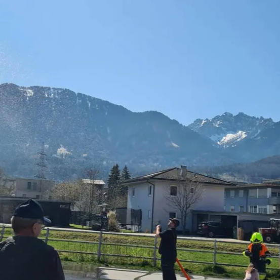 Ein Mann steht auf einem Bürgersteig und sprüht Wasser aus einem Schlauch, während Zuschauer und Kinder zuschauen. Hinter ihm umschließt ein Zaun einen Rasenbereich. Im Hintergrund sind ein Haus und ein Auto zu sehen. Schneebedeckte Berge sind in der Ferne.