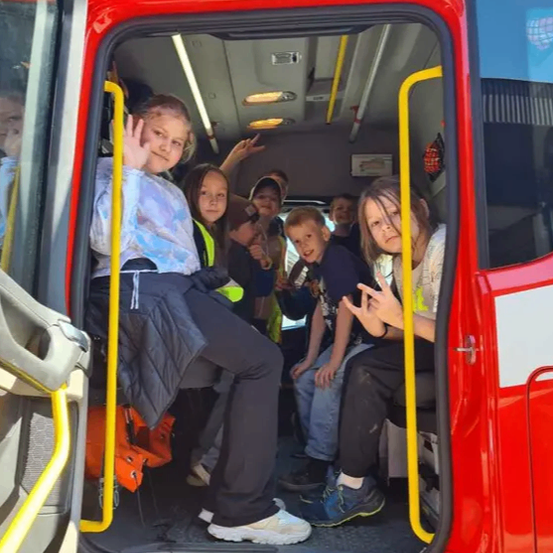 Eine Gruppe von Kindern sitzt in einem roten Feuerwehrauto. Einige tragen Sicherheitswesten, während andere Schuhe anhaben. Die Tür ist offen, und eine Person macht ein Foto.