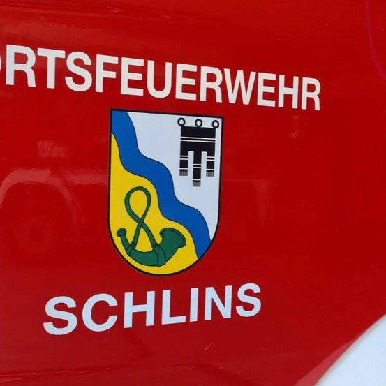 Ein rotes Schild mit weißen Buchstaben zeigt 'ORTSFEUERWEHR SCHLINS'. Das Schild hat einen Wappenschild mit blau-gelbem Hintergrund und ein Logo mit einem Horn und einem schwarzen Symbol.