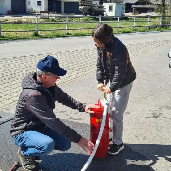 Ein Mann und eine Frau benutzen einen roten Feuerlöscher auf einer Straße mit Häusern und Bergen im Hintergrund.