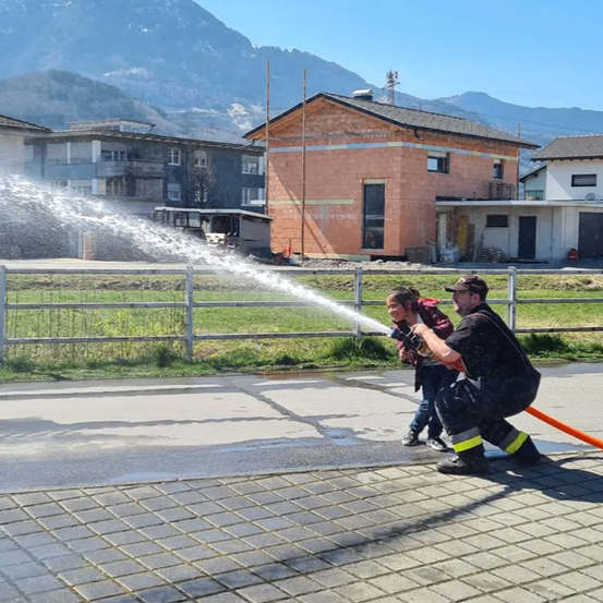 Ein Feuerwehrmann und ein Kind sprühen Wasser aus einem Schlauch vor einem Gebäude. Im Hintergrund befinden sich Häuser und Berge.
