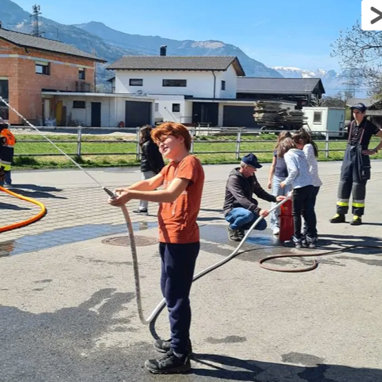Ein Junge hält einen Schlauch, während ein Mann einem Mädchen hilft, einen Feuerlöscher zu bedienen. Sie befinden sich auf einem gepflasterten Bereich mit Häusern und Bergen im Hintergrund.