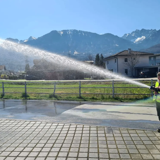 Eine Person benutzt einen Wassersprüher auf einem Ziegelpflaster vor einem Zaun mit einer Bergkette im Hintergrund.