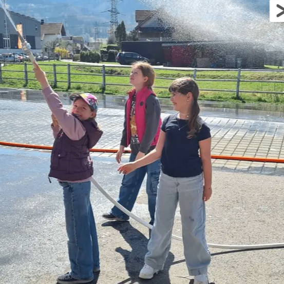 Drei junge Mädchen spielen draußen mit einem Wasserschlauch. Ein Mädchen sprüht Wasser aus dem Schlauch, während die anderen beiden zusehen. Sie stehen auf einem Ziegelpflaster.