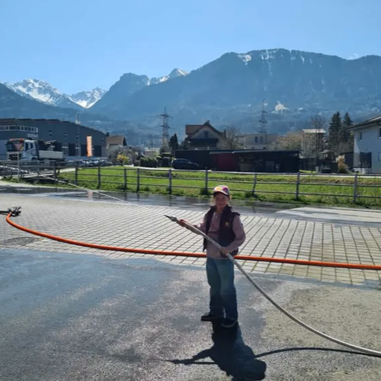 Eine Person benutzt einen langen Schlauch, um Wasser auf einer gepflasterten Straße zu sprühen, mit Bergen im Hintergrund.