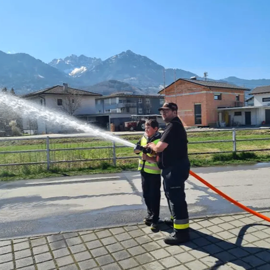 Zwei Männer in Uniformen sprühen Wasser aus einem Schlauch in einer Freiluftumgebung. Ein Grasfeld und Häuser sind im Hintergrund sichtbar, mit Bergen in der Ferne.