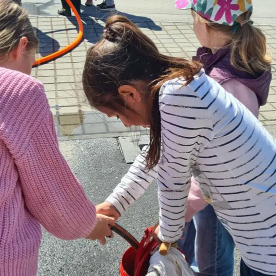 Drei Mädchen füllen einen roten Eimer mit Wasser aus einem Schlauch, wobei ein Mädchen den Griff hält. Sie tragen Freizeitkleidung und es gibt ein verschwommenes Bild von Feuerwehrleuten hinter ihnen.