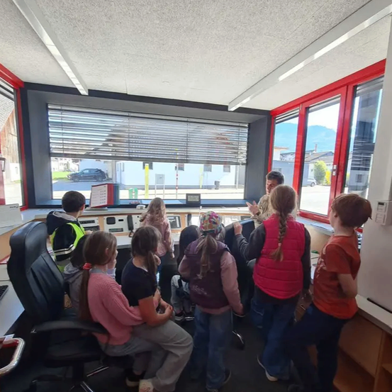 Eine Gruppe von Kindern und zwei Erwachsenen befindet sich in einem Kontrollraum mit großen Fenstern. Draußen sind Autos geparkt und Gebäude sichtbar. Der Raum ist mit Monitoren und Schreibtischen ausgestattet.