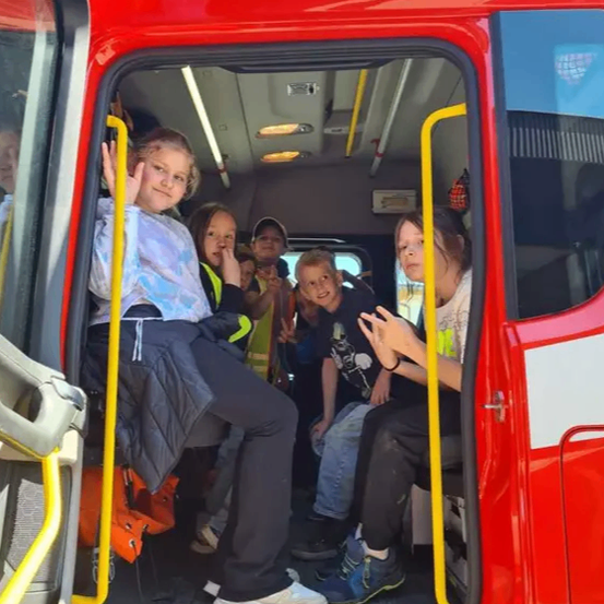 Eine Gruppe von Kindern sitzt in einem roten Notfallfahrzeug und schaut aus der offenen Tür. Ein Kind hält einen gelben Stab und ein anderes winkt.