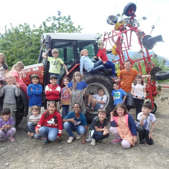Eine Gruppe von Kindern und Erwachsenen posiert für ein Foto vor einem dekorierten Traktor. Einige sitzen auf dem Traktor, andere knien oder stehen. Bäume und ein Berg sind im Hintergrund zu sehen.