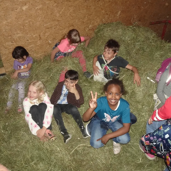 Mehrere Kinder sitzen in einem Heuhaufen. Ein Mädchen mit blonden Haaren und ein Junge in einem blauen Shirt machen Friedenszeichen. Der Raum ist schwach beleuchtet.