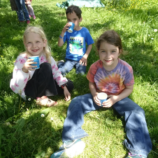 Drei Kinder sitzen auf dem Gras, lächeln und halten Tassen. Ein Mädchen links trägt ein Blumenhemd. Der Junge in der Mitte trägt ein blaues T-Shirt. Ein Mädchen rechts trägt ein Batik-Shirt.
