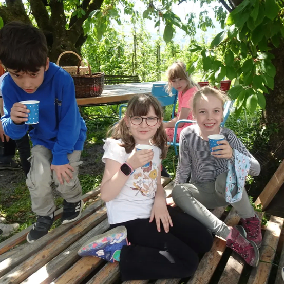 Eine Gruppe von Kindern sitzt auf einer Holzbank im Freien. Zwei Mädchen halten Tassen und ein Junge steht hinter ihnen. Im Hintergrund befinden sich Bäume und ein Tisch.