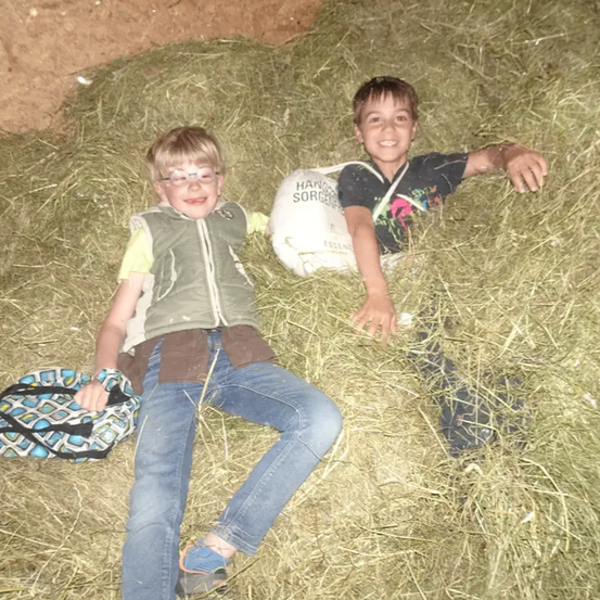 Zwei Jungen liegen in einem Heuboden. Ein Junge trägt eine Brille und eine Weste, der andere hat einen weißen Fußball mit der Aufschrift 'Essence'.