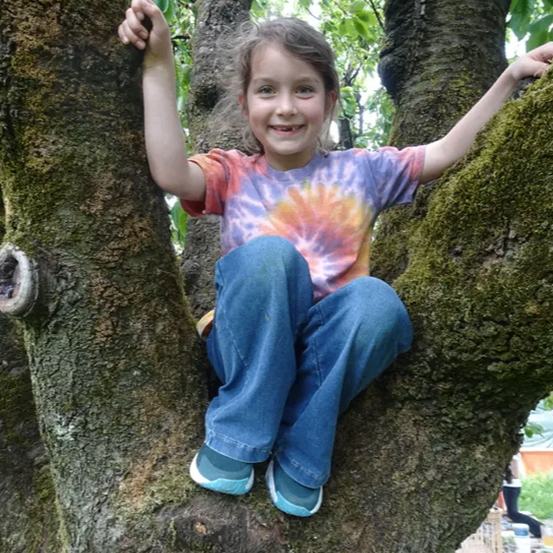 Ein junges Mädchen mit einem Lächeln auf dem Gesicht sitzt auf einem Ast. Sie trägt ein Batik-T-Shirt und blaue Jeans.