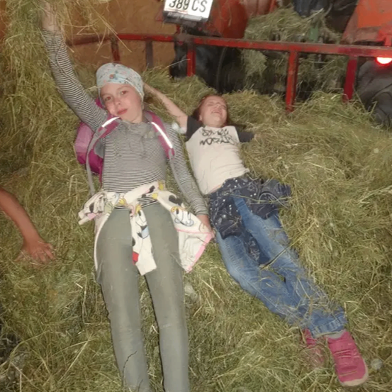 Zwei Mädchen liegen in einem Heuboden mit einem Traktor im Hintergrund, einem roten Zaun und einem Schild mit der Aufschrift 'FK 389 CS'. Sie lächeln und scheinen den Moment zu genießen.