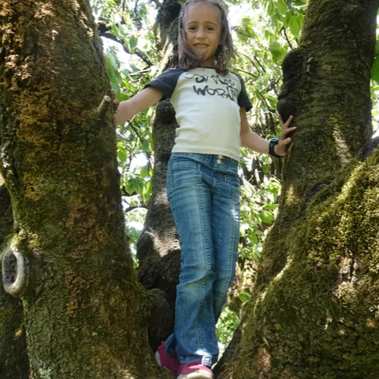Ein junges Mädchen steht zwischen zwei Bäumen, lächelt und posiert für ein Foto. Sie trägt ein weißes T-Shirt mit schwarzem Text und blaue Jeans. Die Bäume haben Moos und grüne Blätter.