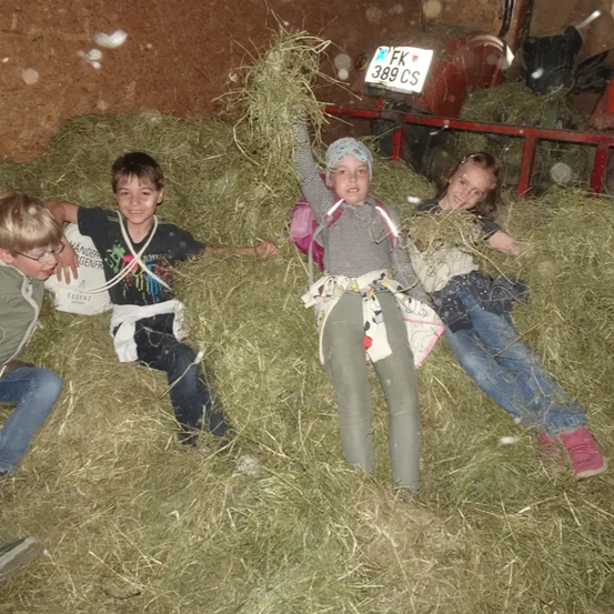 Drei Kinder liegen in einem Heuhaufen in einer Scheune. Eines hebt ein Bündel Heu hoch, ein anderes hat einen Rucksack auf dem Rücken. Im Hintergrund steht eine rote Metallkonstruktion.