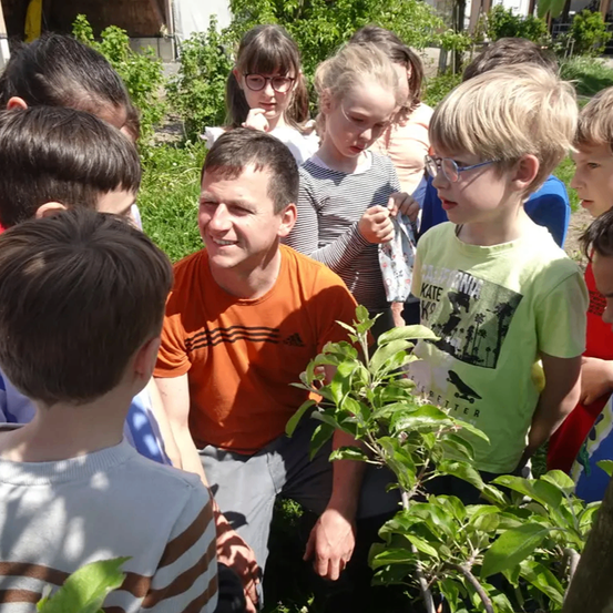 Eine Gruppe von Kindern in einem Garten mit einem lächelnden Mann, der ein oranges T-Shirt trägt und sich hinknien muss, um mit ihnen zu sprechen.