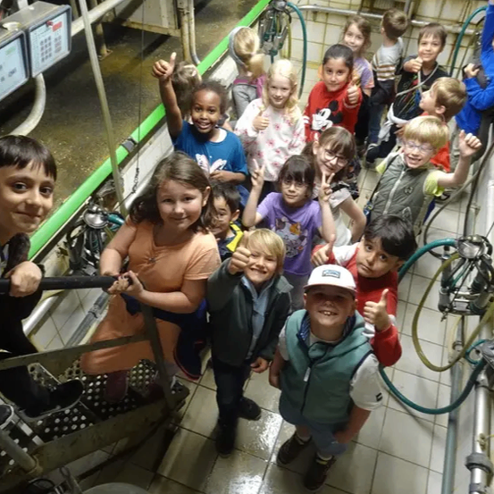 Eine Gruppe von Kindern posiert für ein Foto in einer Melkkammer. Einige lächeln und machen Handzeichen.