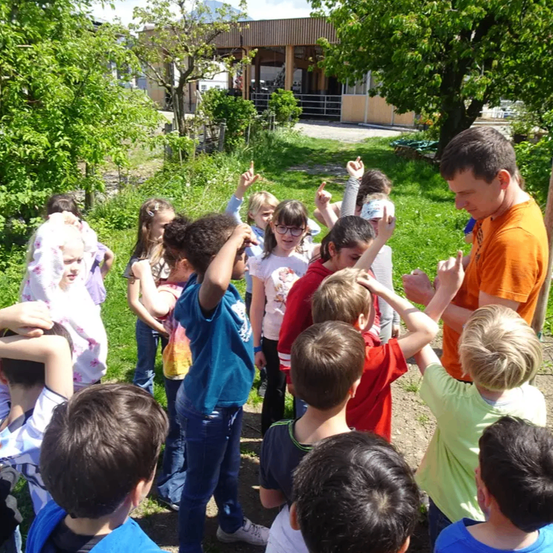 Eine Gruppe von Kindern und ein erwachsener Mann stehen in einer Outdoor-Umgebung. Die Kinder machen verschiedene Handgesten, während der Erwachsene mit ihnen interagiert. Dahinter ist ein Rasenbereich mit Bäumen und einem Gebäude zu sehen.