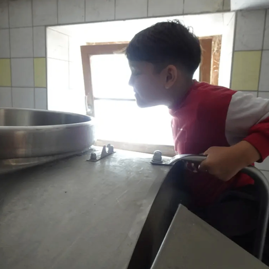 Ein junger Junge steht an einem Waschbecken und hält den Wasserhahn mit der Hand. Er schaut nach unten. Auf dem Tisch steht ein Edelstahltopf.