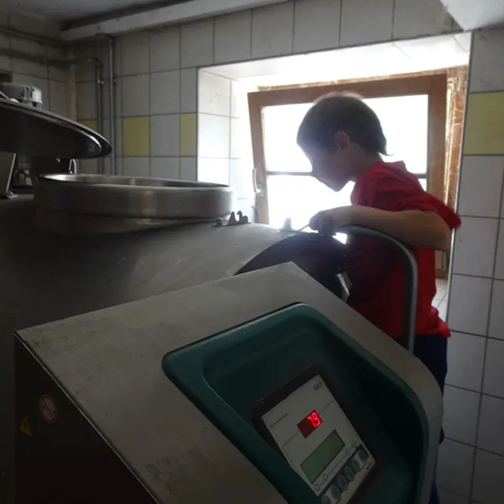 Ein junger Junge in einem roten Shirt bedient eine Edelstahlmaschine in einem gefliesten Raum mit einem hölzernen Fenster.