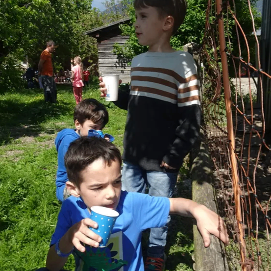 Drei junge Jungen sind in einem Garten. Ein Junge hält einen Becher und ein anderer lehnt sich an einen Zaun. Dahinter steht ein Holzhaus und mehrere Menschen.