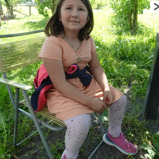 Ein kleines Mädchen mit einer Halskette sitzt auf einem Stuhl im Garten, trägt ein pfirsichfarbenes Kleid und rosa Schuhe. Sie hält ihre Hände zusammen und schaut nach vorne.