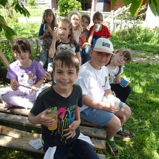 Eine Gruppe von Kindern sitzt draußen auf einer Holzbank, einige halten Tassen. Sie lächeln und scheinen für ein Foto zu posieren. Sie sind von Grün umgeben.