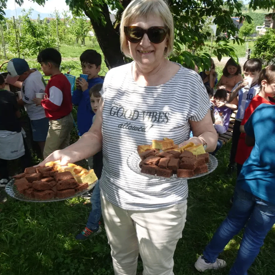 Eine Frau mit einem gestreiften T-Shirt mit der Aufschrift 'GOOD VIBES ALWAYS' hält zwei Teller mit Brownies in einem Park mit Kindern.