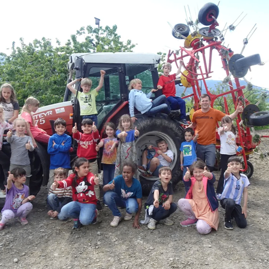 Eine Gruppe von Kindern und zwei Erwachsenen posiert für ein Foto vor einem roten Traktor mit einem Reifen oben drauf. Die Kinder lächeln und geben Daumen hoch.