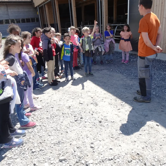 Eine Gruppe von Kindern in verschiedenen farbigen Kleidungsstücken steht in einer Reihe und blickt auf einen Mann in einem orangefarbenen T-Shirt. Er scheint sie anzusprechen. Sie befinden sich in einem Kiesbereich mit einem Gebäude im Hintergrund.