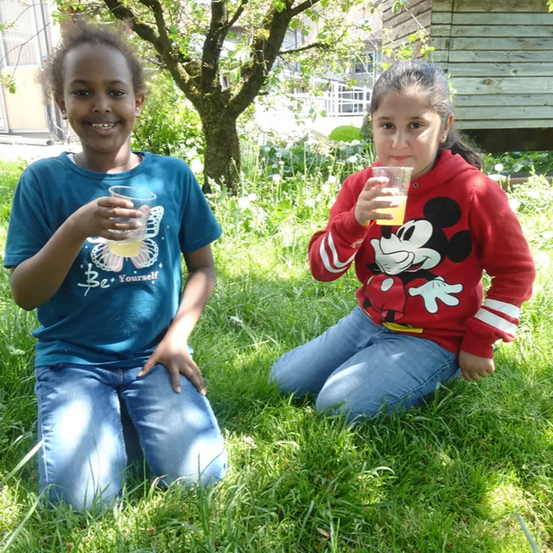 Zwei Kinder sitzen auf einer Rasenfläche. Ein Mädchen in einem Mickey-Mouse-Hoodie trinkt Orangensaft. Ein Junge in einem blauen T-Shirt lächelt und hält einen Glas mit Saft. Ein Baum und ein Holzhaus sind im Hintergrund.