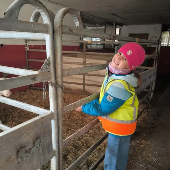 Ein Mädchen in einer Warnweste steht in einer Scheune, lächelt und hält sich an einem Metalltor fest. Heu ist auf dem Boden sichtbar.