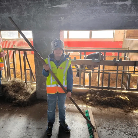 Ein Junge in einem gelben Weste und Mütze steht in einer Scheune und hält einen Besen. Hinter ihm ist durch ein Metallgitter eine Kuh zu sehen. Heu liegt auf dem Boden.