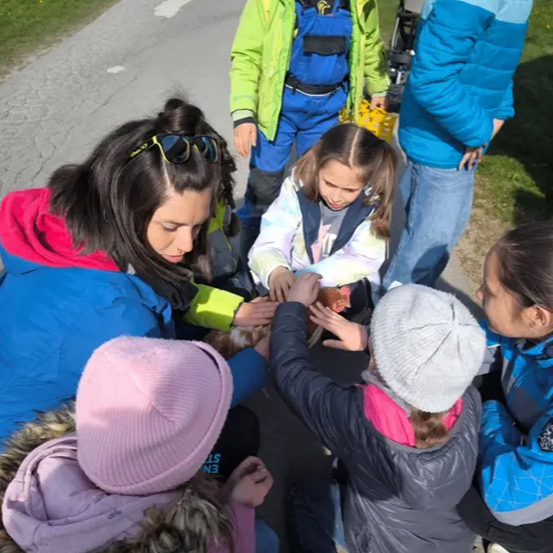 Eine Gruppe von Kindern und Erwachsenen versammelt sich auf einem Weg und betrachtet ein Huhn. Sie tragen alle Winterkleidung.