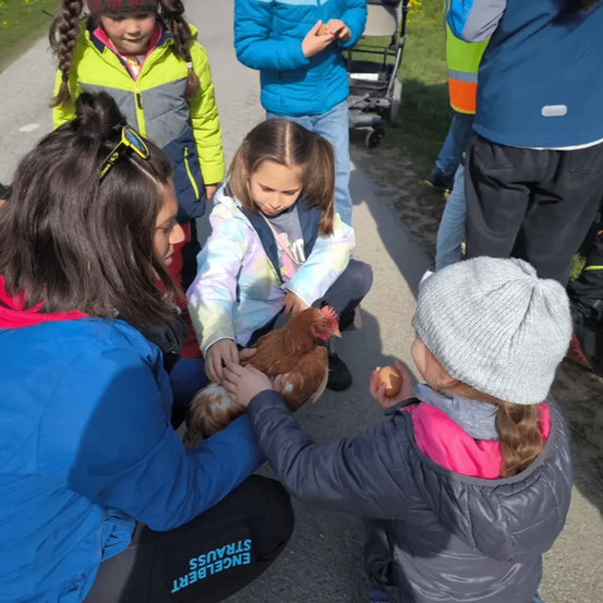 Eine Gruppe von Kindern versammelt sich um eine Frau, die ein Huhn hält. Die Kinder beobachten aufmerksam, und eines hält ein Ei.