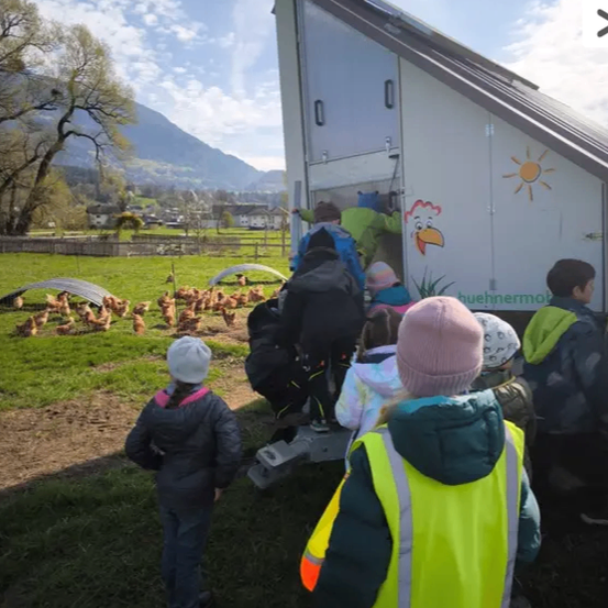 Kinder versammeln sich um einen weißen Schuppen mit einem Hahn an der Seite. Einige tragen Rucksäcke und andere tragen Mützen. Sie beobachten, wie eine Person den Schuppen betritt.